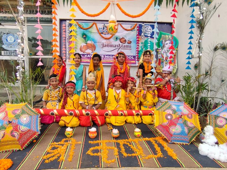 Children in yellow and orange costumes pose for Janmashtami at-school celebration with marigold rangoli and bright umbrellas in the background.